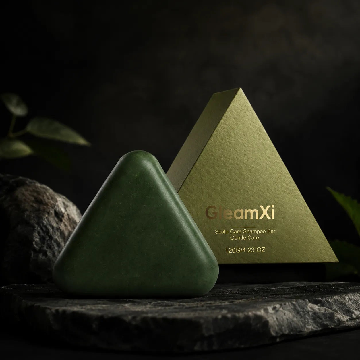 A dark green triangular shampoo bar sits in front of a metallic gold pyramid-shaped box labeled 'GleamXi Scalp Care Shampoo Bar Gentle Care' on a dark stone surface against a dark background.